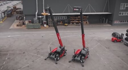 Two red Magni telehandlers with tall black extended booms parked side by side in an industrial yard outside a warehouse site