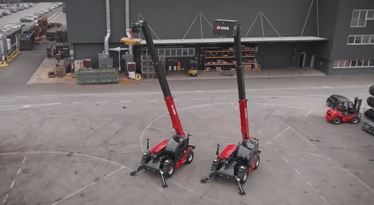 Two red Magni telehandlers with tall black extended booms parked side by side in an industrial yard outside a warehouse site