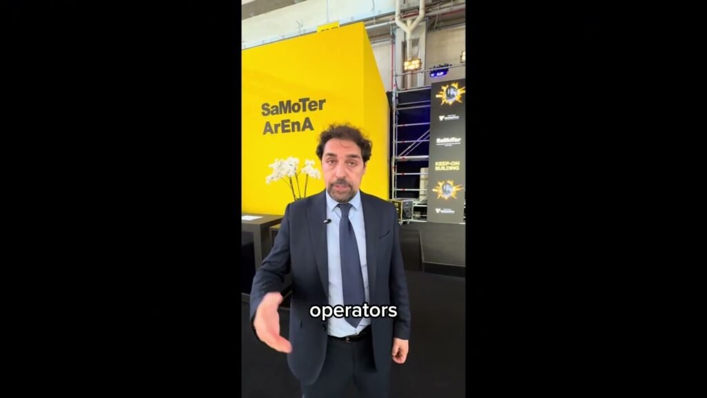Man in a dark suit and tie speaks at a convention booth, with a bright yellow backdrop reading 'SaMoTer Arena'.