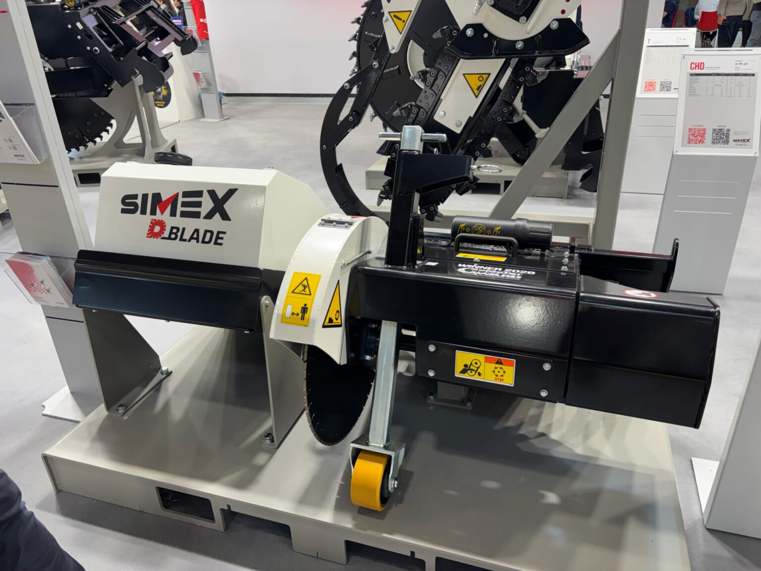 simex-d-bladejpg - Machines Wiki Industrial SIMEX Blade machine on display at a trade show, showing warning stickers, yellow roller and dark metal framework used for cutting or shredding material