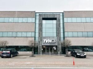 Office building entrance with a tall glass-and-stone facade, TVH logo above the doors, two leafless trees, and parked cars in front of a paved area.