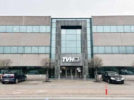 tvhjpeg - Machines Wiki Office building entrance with a tall glass-and-stone facade, TVH logo above the doors, two leafless trees, and parked cars in front of a paved area.