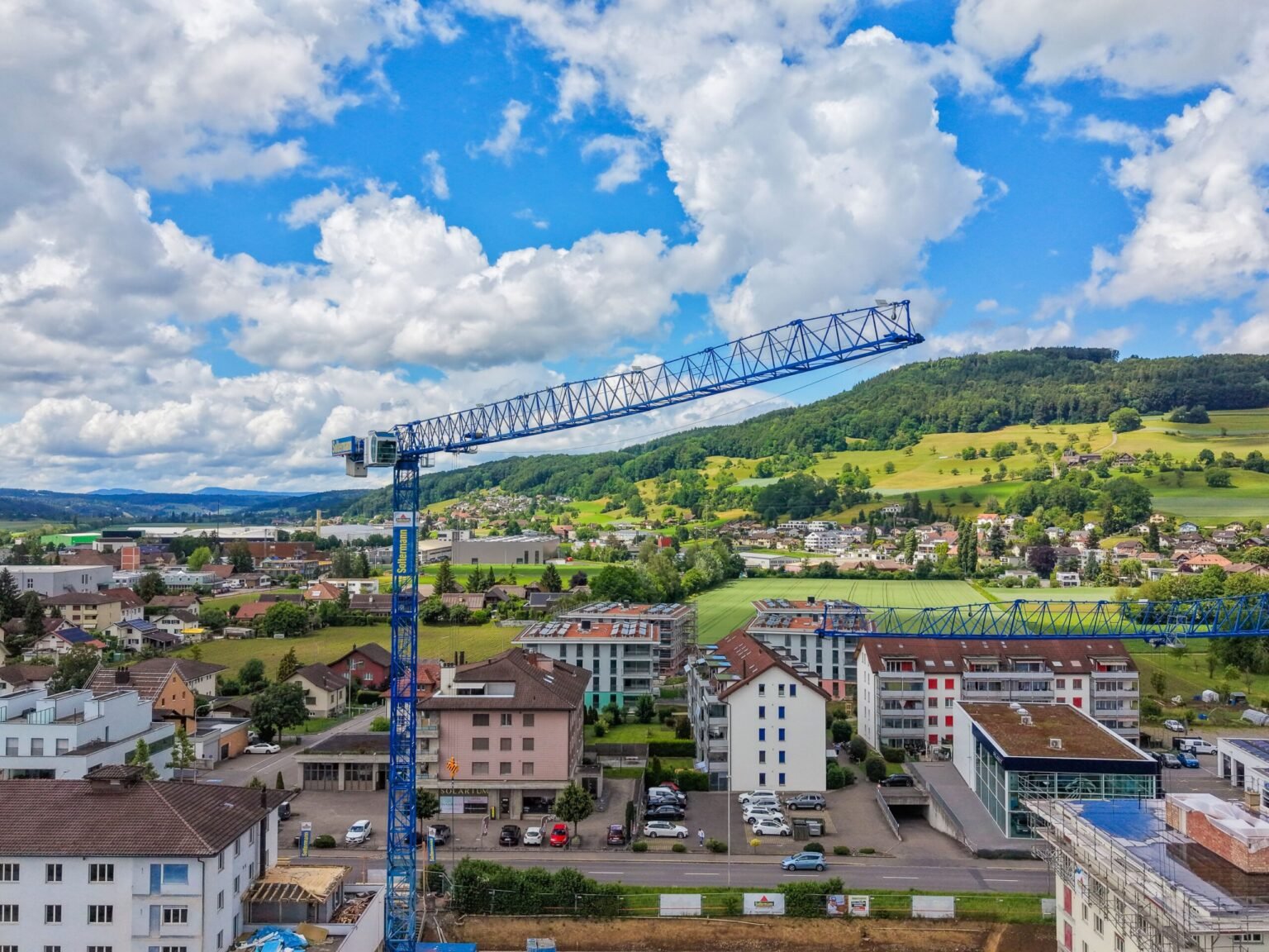 Blue construction crane stretching over a residential area with rolling green hills and blue sky in the background.