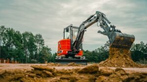 Orange excavator on a dirt lot scooping soil with its bucket, trees in the background under a cloudy sky