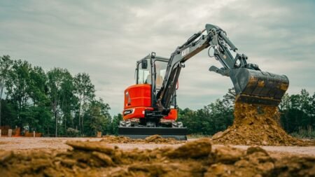 Orange excavator on a dirt lot scooping soil with its bucket, trees in the background under a cloudy sky
