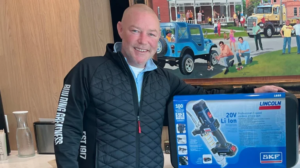 Smiling man in a quilted black jacket stands beside a blue box for a 20V cordless grease gun on display in a showroom.