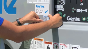 Close-up of a person's hands attaching a label to a Genie industrial lift, with safety stickers and a control panel visible in the background.