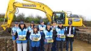 Group of ten students in blue safety vests posing in front of a yellow Komatsu excavator at a construction site, each holding a boxed item.