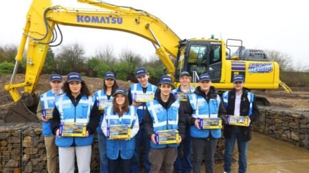 Group of ten students in blue safety vests posing in front of a yellow Komatsu excavator at a construction site, each holding a boxed item.