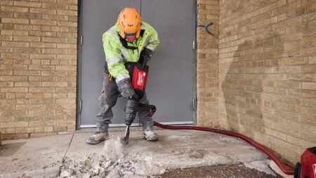 Construction worker in safety gear breaking concrete with a jackhammer beside a doorway in an exterior area.