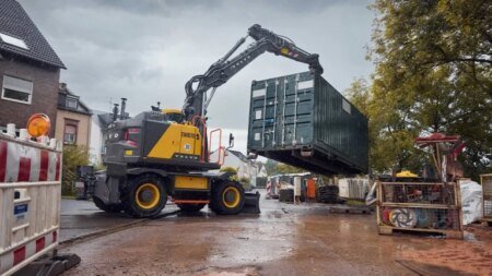 A yellow and black excavator lifting a large green container at a construction site, with barriers and equipment around.