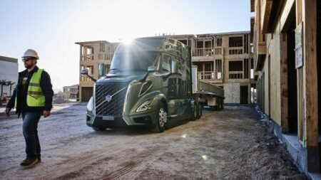 1780939558_63774_en_e3c0c_65909_volvo-trucks-remote-updates-2jpg - Machines Wiki Construction site with a green semi-truck and trailer; a worker in a hard hat and reflective vest walks beside it, wooden building frames in the background.