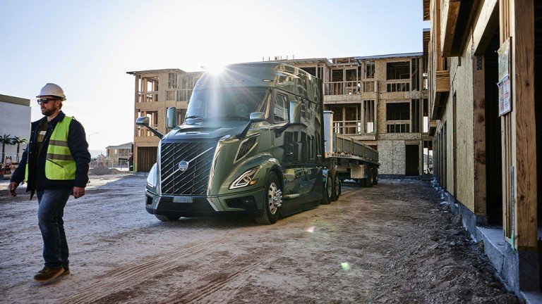 1780939558_63774_en_e3c0c_65909_volvo-trucks-remote-updates-2jpg - Machines Wiki Construction site with a green semi-truck and trailer; a worker in a hard hat and reflective vest walks beside it, wooden building frames in the background.