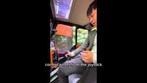1781013404_maxresdefaultjpg - Machines Wiki Person in a suit sits inside the cab of an orange construction machine, holding a joystick to operate the controls.