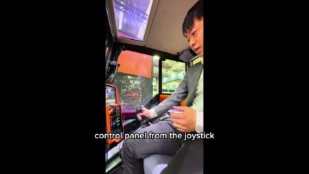 1781013404_maxresdefaultjpg - Machines Wiki Person in a suit sits inside the cab of an orange construction machine, holding a joystick to operate the controls.