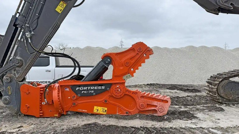 Easily transition from primary demolition to secondary sizing with ShearCore’s new modular concrete processor Orange Fortress FC-75 mulcher attachment on an excavator, set on a gravel surface with a pile of material in the background and tracks visible to the right.