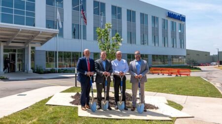 Four men in suits with shovels pose for a groundbreaking ceremony outside a modern office building with glass windows and flags nearby.