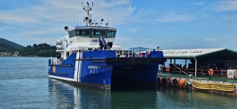 Blue-and-white vessel named KWINDS 1 docked at a harbor quay with a covered dock structure and calm water nearby.