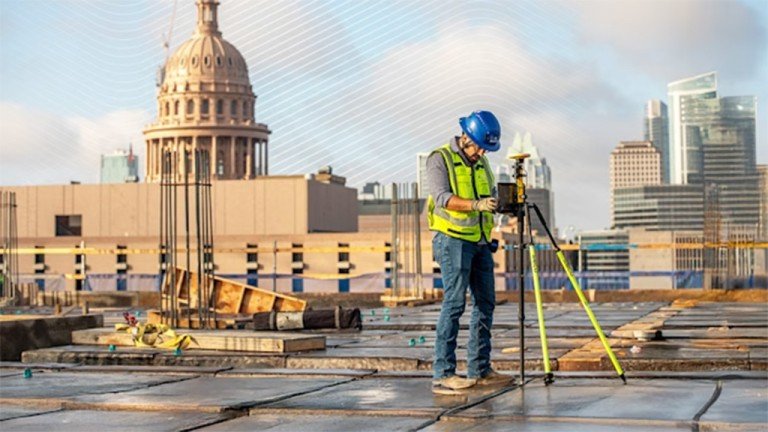 Construction worker in a blue hard hat and safety vest setting up a surveying tripod on a rooftop, with a city skyline in the background.