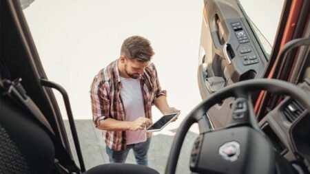 1781729206_63835_en_72a99_65979_geotab-work-driverjpg - Machines Wiki Man in plaid shirt stands outside a truck cab, using a tablet to inspect or navigate.