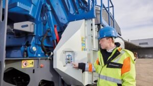 Construction worker in a hard hat and high-visibility jacket operates a large blue industrial machine outdoors, pressing a control panel button.
