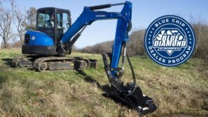 1781740260_63834_en_8e496_65978_blue-diamond-brush-cutter-blue-chip-dealerjpg - Machines Wiki Blue compact excavator with hydraulic arm and bucket on a grassy field, featuring a Blue Diamond Attachments program badge on the right side.