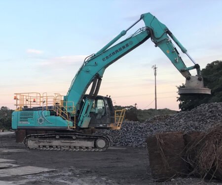 Teal hydraulic excavator on tracks extending its long arm with a clamshell bucket, grabbing rocks at a gravel yard.