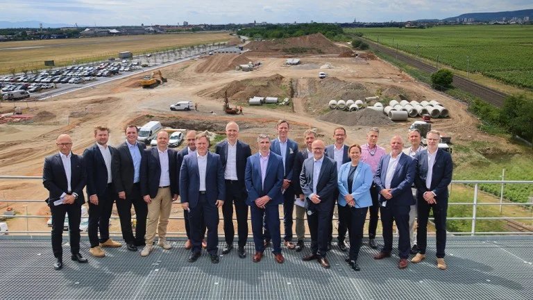 Group of professionals in suits standing on a platform overlooking a construction site with earthworks, equipment, and stacked pipes.