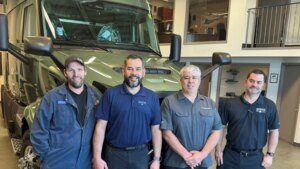 Four technicians pose in front of a large green service van inside a workshop.