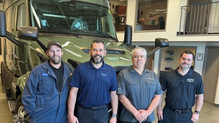 Four technicians pose in front of a large green service van inside a workshop.