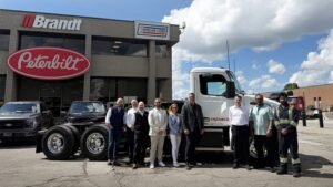 1782238987_63900_en_b3bc0_66034_brandt-peterbilt-548ev-truckjpg - Machines Wiki Twelve people pose in front of a white Peterbilt semi-truck outside a dealership with Brandt and Trans-Corp signs; several black trucks parked nearby.