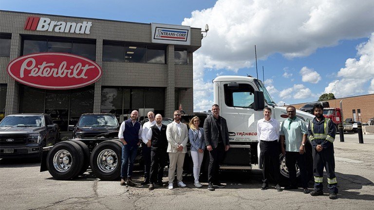 Twelve people pose in front of a white Peterbilt semi-truck outside a dealership with Brandt and Trans-Corp signs; several black trucks parked nearby.