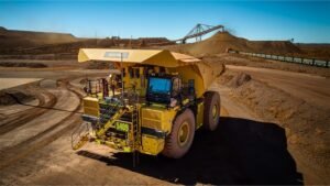 Large yellow mining dump truck at a quarry with dirt piles and a conveyor in the background.
