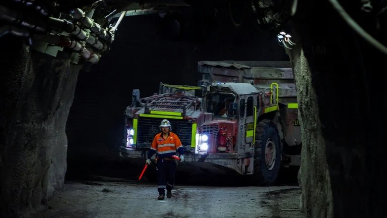 Canada’s 40 Under 40 for industrial leaders returns to redefine the industry Worker in orange safety vest and helmet directing a large dump truck through a dark underground mine passage.