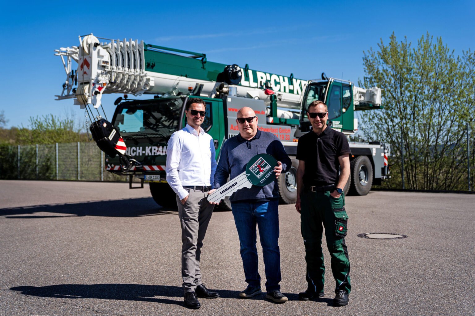 1782297917_liebherr-ltm-1090-4-2-ullrich-krane-300dpi-scaledjpg - Machines Wiki Three men pose in front of a large green mobile crane, holding a oversized ceremonial key.