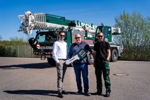 1782297917_liebherr-ltm-1090-4-2-ullrich-krane-300dpi-scaledjpg - Machines Wiki Three men pose in front of a large green mobile crane, holding a oversized ceremonial key.