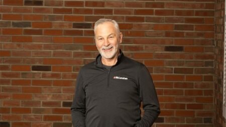 Smiling middle-aged man with gray hair and beard in a black polo with a McLanahan logo, standing against a brick wall.