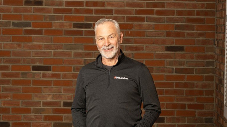 Smiling middle-aged man with gray hair and beard in a black polo with a McLanahan logo, standing against a brick wall.
