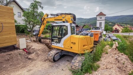 Yellow Liebherr excavator on a dirt construction site, digging a trench beside a wooden shed with a rural hillside backdrop.