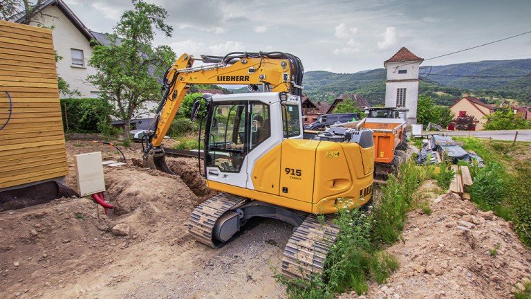 Yellow Liebherr excavator on a dirt construction site, digging a trench beside a wooden shed with a rural hillside backdrop.