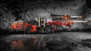 Orange underground mining drill rig with extendable booms, parked in a dark cavern.