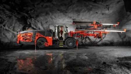 Orange underground mining drill rig with extendable booms, parked in a dark cavern.