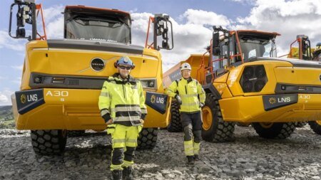 Two construction workers in yellow high-visibility gear stand in front of large yellow Volvo A30 dump trucks on a rocky quarry with a blue sky.