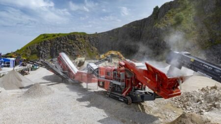Industrial rock crushing site with red machinery and conveyors moving gravel on a dusty quarry floor near rocky cliffs.