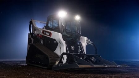 Bobcat skid-steer loader with a front bucket on a dirt surface at night, headlights on illuminate the site