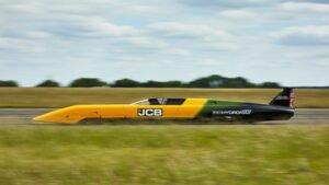 Side view of a bright yellow and black land-speed racing car (JCB Hydra Max) speeding along a track on a clear day with a low, pointed nose and an open cockpit.