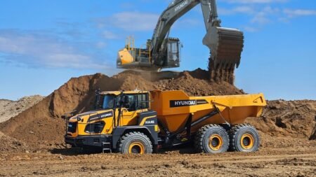 Yellow Hyundai excavator dumps dirt onto a large mound while a Hyundai articulated dump truck waits nearby at a construction site.