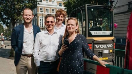 Four adults stand together on a sunny city street beside a Volvo electric construction machine, smiling at the camera.