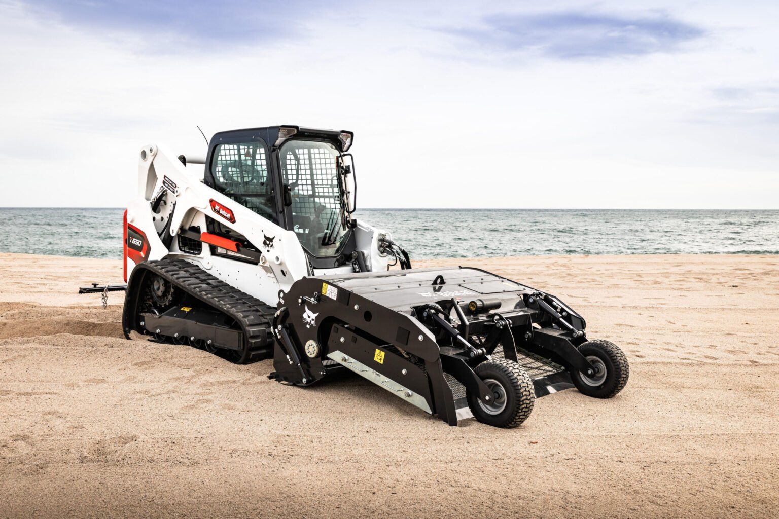 White Bobcat skid-steer loader on a sandy beach with the sea and blue sky in the background for context and scale.