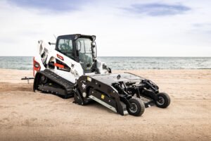 White Bobcat skid-steer loader on a sandy beach with the sea and blue sky in the background for context and scale.